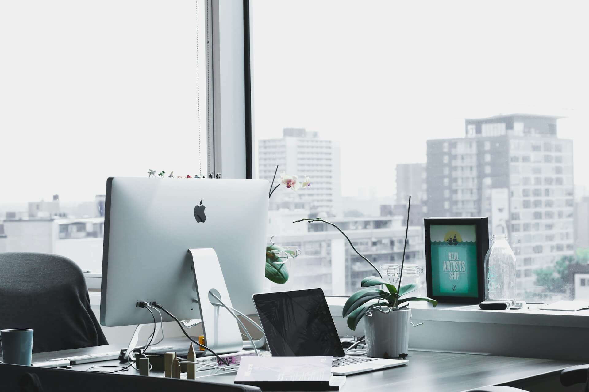 Free Zone Business Setup - Modern office desk with iMac, laptop, and plants by a window overlooking city buildings.