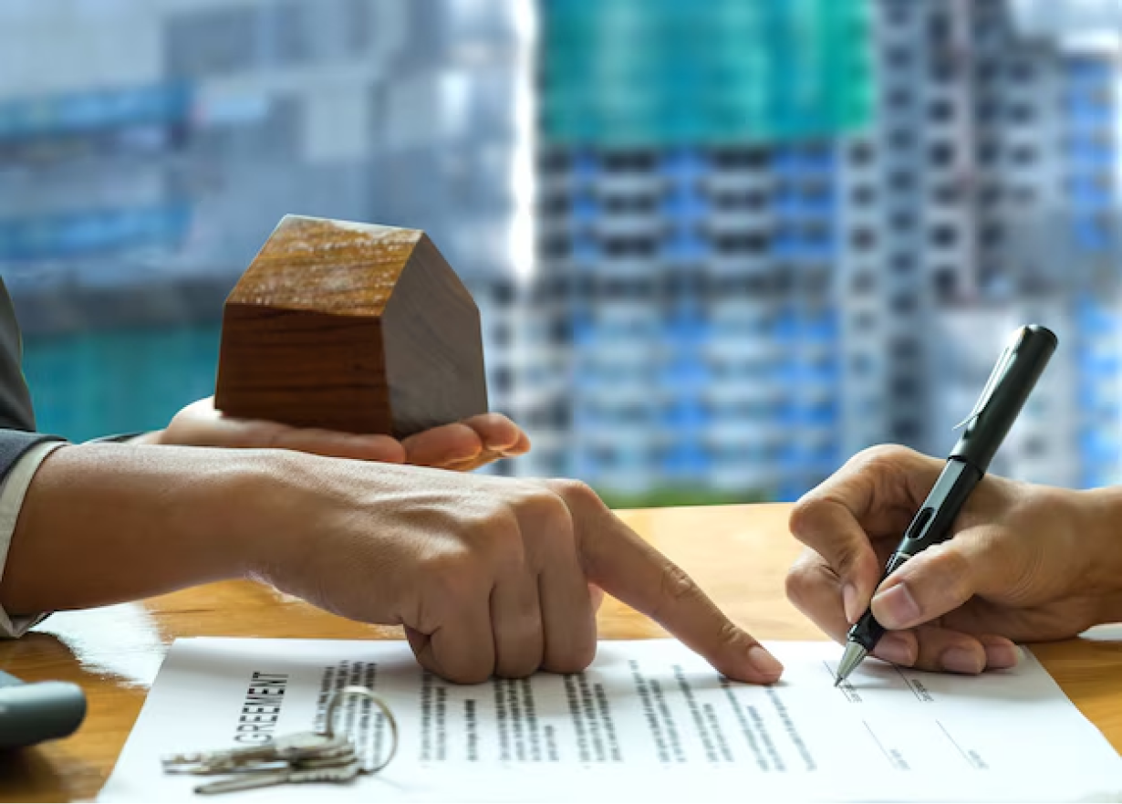 Real Estate Licence in Dubai - Two hands signing a contract with a wooden house model and keys on the table, blurred cityscape background.